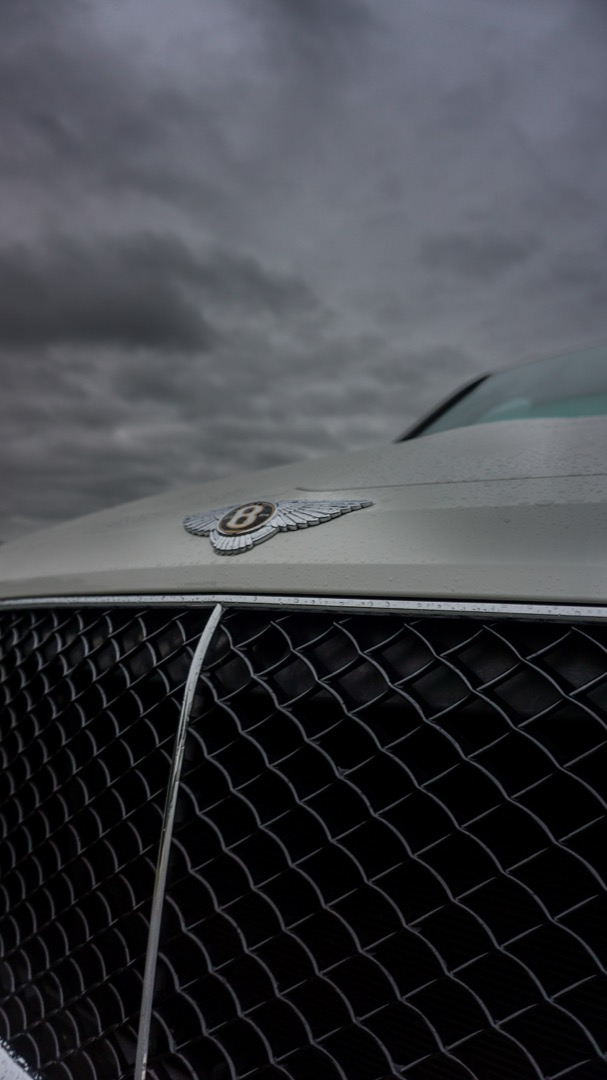 Bentley winged-B emblem on a Bentayga grille against an overcast sky.